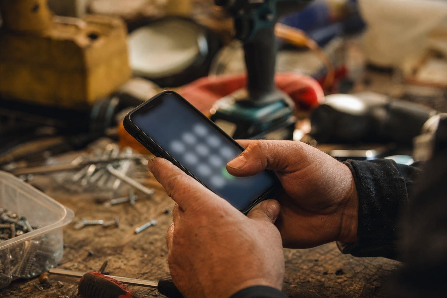 Close-up of a tradesperson's weathered hands holding a smartphone with a thumb on the dial pad over a cluttered workbench of tools and screws.